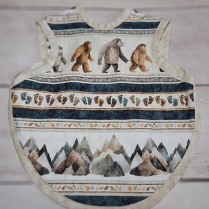 May include: A baby bib featuring a repeating pattern of brown and grey Bigfoot creatures, a row of colourful footprints, and a landscape of mountain peaks. The bib has a cream-coloured trim and a sleeveless design with a rounded neckline.