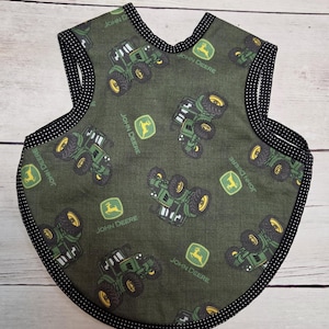 May include: A sleeveless bib featuring an olive green fabric with a repeating pattern of green John Deere tractors and yellow John Deere logos. The edges of the bib are finished with a black fabric trim that has a white polka dot pattern.