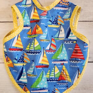 Può includere: Un bavaglino per bambini con sfondo blu raffigurante colorate barche a vela con vele a righe e fantasia. Il bavaglino è rifinito con tessuto giallo e presenta un motivo a pois gialli sul bordo.