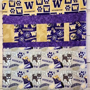 May include: A purple, gold, and gray patchwork quilt with a repeating pattern of the University of Washington Huskies logo. The quilt features the words "Washington Huskies" and "Purple Reign".
