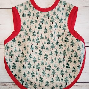 May include: A baby bib with a festive Christmas tree pattern. The bib features a repeating pattern of green Christmas trees with star toppers on a light background. The edges are trimmed with a bright red fabric.