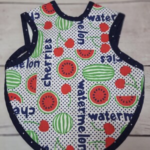 May include: A sleeveless baby bib with a white background featuring a black polka dot pattern, red cherries, green striped watermelons, and red watermelon slices. The text "cherries" and "watermelon" is printed in blue, with dark blue trim.