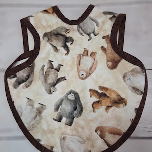 May include: A sleeveless bib featuring a cream-coloured background with a repeating pattern of various brown, grey, and white yeti or sasquatch creatures. The bib is finished with a dark brown fabric trim along the neck, armholes, and outer edges.