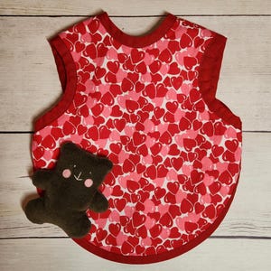 May include: A red sleeveless bib with a white background and a pattern of red and pink hearts. A brown teddy bear is sitting on the bib.
