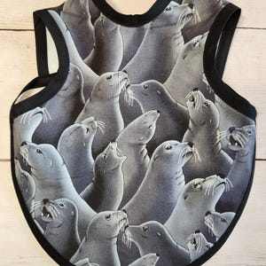May include: A baby bib with a repeating pattern of grey seals with white whiskers. The bib has a black trim and a curved shape. The seals are in various poses, some with their mouths open.