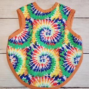 May include: A tie-dye baby bib with a brown trim. The bib has a vibrant spiral pattern in green, yellow, red, blue, and orange. The bib is designed to protect clothing during mealtime.