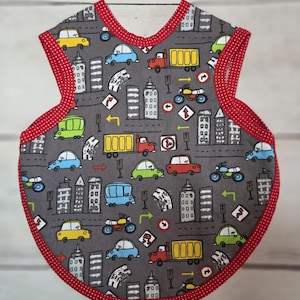 May include: A sleeveless baby bib with a grey background featuring a repeating pattern of colourful cars, trucks, motorcycles, and city buildings. The bib is finished with a red trim that has a white polka dot pattern along the edges.