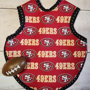 May include: A red baby bib featuring the San Francisco 49ers logo and text. The bib is edged with black polka dot fabric. A toy football is also present. The bib is approximately 25 cm wide.