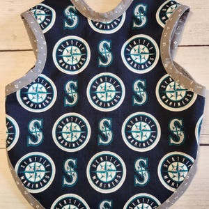 May include: A navy blue baby bib with a repeating pattern of the Seattle Mariners logo and the letter "S". The bib has a grey trim with small white polka dots.