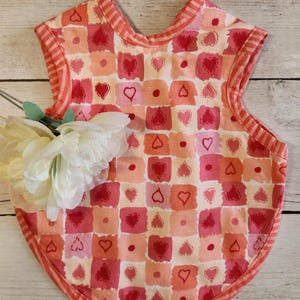 May include: A baby bib with a heart pattern in shades of pink and peach. The bib has a rounded bottom and a striped trim. A white flower is placed on the left side of the bib.