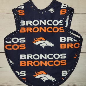 May include: A navy blue baby bib with the word "BRONCOS" in white and orange, and the team's logo. The bib has a rounded bottom and white polka dots along the edges.