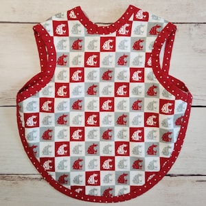 May include: A baby bib with a red and white checkered pattern featuring a cougar logo. The bib has a red polka dot trim and is designed to protect clothing during mealtime. The bib is made of fabric.