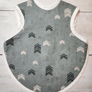 May include: A grey baby bib with a white trim and a pattern of black and white arrows. The bib has a rounded shape and is made of fabric.