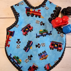May include: A baby bib with a light blue background featuring colorful train cars and railroad crossing signs. The bib has a black polka dot trim and is next to some red, black, and blue toy blocks.