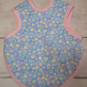 Könnte beinhalten: Ein ärmelloses Lätzchen mit hellblauem Hintergrund und einem Muster aus bunten Kreisen in Rosa, Gelb, Violett und Türkis. Das Lätzchen ist mit einer hellrosa Einfassung versehen, die ein dezentes weißes Punktemuster an den Rändern aufweist.