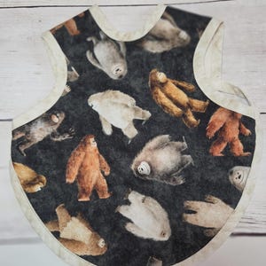 May include: A baby bib featuring a dark grey background with a repeating pattern of various brown, white, and grey yeti or sasquatch figures. The bib has a rounded shape with a cream-coloured fabric trim along the edges and a neck opening.