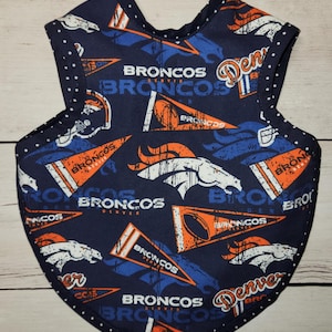 May include: A navy blue baby bib with a repeating pattern of Denver Broncos logos, helmets, and pennants in orange, white, and blue. The bib has a white polka dot trim.