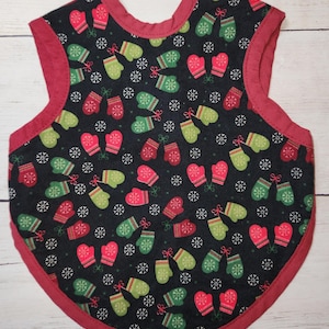 May include: A baby bib with a festive winter design. The bib has a black background patterned with red and green mittens and white snowflakes. The edges are trimmed with a solid red fabric.