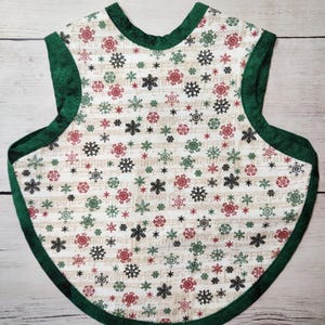 May include: A festive bib with a cream background, decorated with red, green, and black snowflakes. The bib is edged with dark green fabric. The design incorporates musical notes, adding a unique touch. The bib is designed for a baby.
