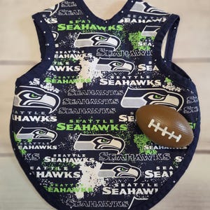 Può includere: Una bavaglina per neonati blu navy con un motivo ripetuto del logo e della scritta Seattle Seahawks in bianco e verde neon, con un piccolo pallone da football marrone con lacci bianchi appoggiato sulla superficie del tessuto.