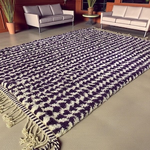 Puede incluir: Una alfombra rectangular con un patrón geométrico en tonos morados y crema. La alfombra tiene una superficie texturizada y está rematada con flecos en los bordes. La alfombra está colocada sobre un suelo de color neutro.