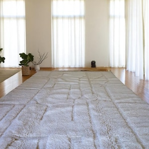 Op de afbeelding: Een ruime kamer met een groot, lichtgrijs rechthoekig tapijt met een geometrisch patroon. Het tapijt ligt op een houten vloer, met grote ramen met witte gordijnen. Potplanten voegen een vleugje groen toe aan de ruimte.