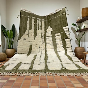 Puede incluir: Una alfombra grande, de color verde oliva y crema, con un patrón geométrico. La alfombra tiene un aspecto texturizado y peludo y se exhibe en una habitación bien iluminada con plantas y un suelo de baldosas. La alfombra tiene borlas en los bordes.