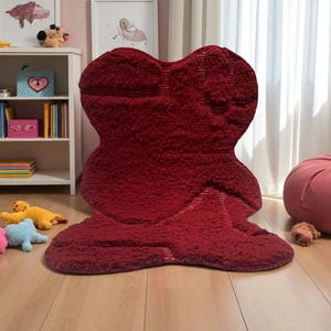 May include: A large, plush, red rug in an abstract shape dominates the room. The rug has a textured surface and is placed on a wooden floor. Additional plush toys in various colors are scattered around the rug, suggesting a children's room.