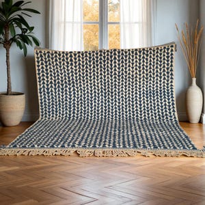 May include: A large, rectangular rug with a repeating pattern of navy blue arrow shapes on a cream background. The rug has a fringed edge and is displayed indoors on a hardwood floor. A potted plant and a vase are visible.