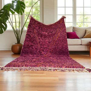 Puede incluir: Una alfombra grande y texturizada en tonos de rosa intenso y morado, con un patrón geométrico dorado. La alfombra tiene un borde con flecos en crema y naranja. La alfombra se exhibe en una habitación bien iluminada con un sofá y una gran planta en maceta.