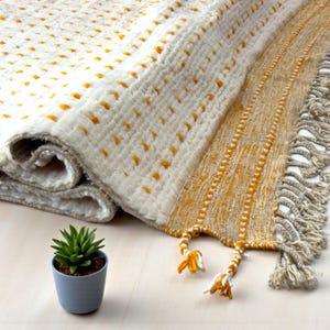 May include: A folded white and yellow patterned rug with a fringed edge. The rug has a textured design with yellow dots and stripes. A small blue pot with a green plant is in the foreground.