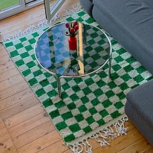 Puede incluir: Un sofá seccional gris con una almohada blanca con un diseño de palmera se encuentra en una sala de estar con una alfombra a cuadros verde y blanca. Una mesa de café de vidrio con una base cromada se encuentra sobre la alfombra. Un jarrón con flores rosas se encuentra sobre la mesa.