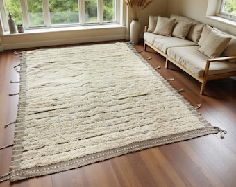 May include: A cream-coloured rectangular rug with a textured, striped pattern. The rug features a border with a dotted design and tassels on the short edges. It is displayed on a wooden floor in a living room.