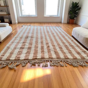 Elegant handwoven Moroccan rug with vertical stripes in Cameo Rose and soft white. Perfect for boho, coastal, minimalist, or Japandi-inspired spaces. Made from natural wool, this artisan piece blends texture and comfort for your living room, bedroom, or entryway. A modern pastel rug with warm earthy tones and timeless Amazigh character. Custom sizes available.