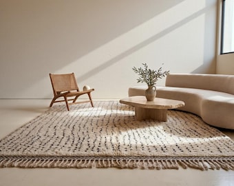 May include: A cream-colored rug with a black dotted pattern and fringed edges. The rug is placed in a room with a wooden chair, a coffee table, and a curved sofa. The room has a neutral color palette.