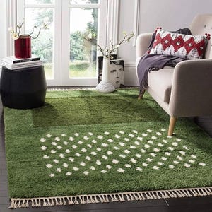 A plush handwoven Moroccan Berber rug in a rich moss green. The thick wool carpet features a solid dark green band near the top and a lighter mossy green base beautifully adorned with small, cream-colored geometric diamond motifs at the bottom. It is draped elegantly against a white painted brick wall next to a gold accent table with trailing ivy, perfectly capturing a warm, earthy, and organic modern interior design aesthetic.