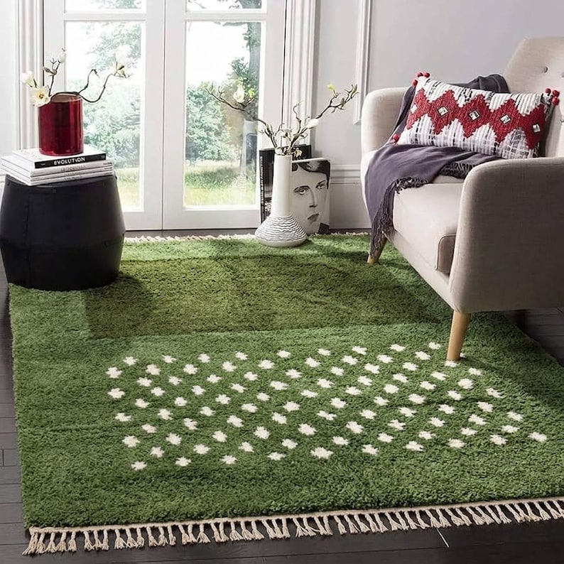 A plush handwoven Moroccan Berber rug in a rich moss green. The thick wool carpet features a solid dark green band near the top and a lighter mossy green base beautifully adorned with small, cream-colored geometric diamond motifs at the bottom. It is draped elegantly against a white painted brick wall next to a gold accent table with trailing ivy, perfectly capturing a warm, earthy, and organic modern interior design aesthetic.