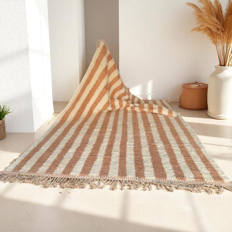 Elegant handwoven Moroccan rug with vertical stripes in Cameo Rose and soft white. Perfect for boho, coastal, minimalist, or Japandi-inspired spaces. Made from natural wool, this artisan piece blends texture and comfort for your living room, bedroom, or entryway. A modern pastel rug with warm earthy tones and timeless Amazigh character. Custom sizes available.