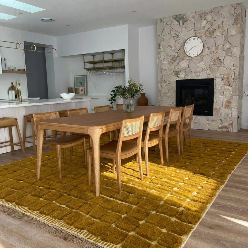 Large handmade Moroccan high-pile shag rug in saturated Thai curry mustard-gold, displayed vertically against a white brick wall. Surface features a tone-on-tone square grid pattern formed through pile variation — raised mustard squares separated by recessed ivory cream channel lines across the entire field. Dense, plush, curly wool pile texture throughout. Knotted braided ivory fringe along the top hem edge and long hand-tied ivory fringe along the bottom edge.