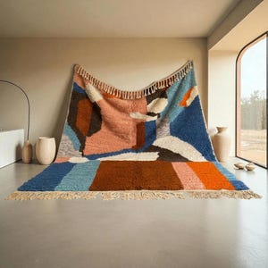 Puede incluir: Una alfombra grande y colorida con un diseño geométrico. La alfombra presenta una mezcla de colores que incluyen azul, naranja, marrón y rosa. Tiene un borde con flecos y se exhibe contra una pared neutra.