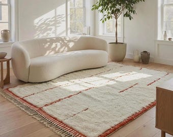 May include: A rectangular area rug featuring a thick, cream-coloured wool pile with horizontal, rust-coloured linear patterns and a fringed edge. The rug is positioned on a light-toned wooden floor in a room with a curved, cream-coloured sofa.