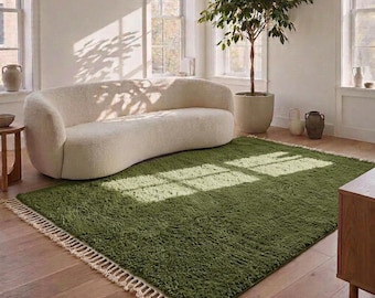 Può includere: Un tappeto rettangolare di colore verde oliva intenso caratterizzato da una consistenza a pelo lungo e spesso, con dettagli a frange annodate color crema lungo i due bordi più corti, posizionato su un pavimento in legno chiaro.
