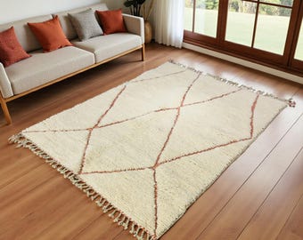 May include: Cream-coloured rectangular rug featuring a geometric diamond pattern in a reddish-brown hue. The rug has a textured surface and fringe detailing along the edges. The rug is placed on a hardwood floor.