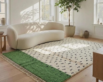 May include: A rectangular area rug featuring a cream-coloured field with a repeating pattern of small black marks and a solid green band on one end. The rug is finished with cream-coloured fringe along the edges and has a thick, textured pile.