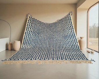 May include: A large rectangular area rug featuring a repeating dark blue geometric pattern on a cream-colored background. The rug has a textured surface with visible tufted details and a decorative fringe along the bottom edge.