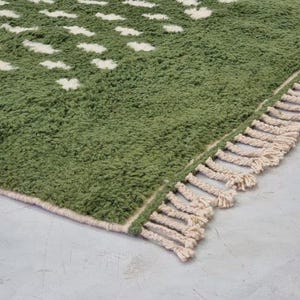A plush handwoven Moroccan Berber rug in a rich moss green. The thick wool carpet features a solid dark green band near the top and a lighter mossy green base beautifully adorned with small, cream-colored geometric diamond motifs at the bottom. It is draped elegantly against a white painted brick wall next to a gold accent table with trailing ivy, perfectly capturing a warm, earthy, and organic modern interior design aesthetic.
