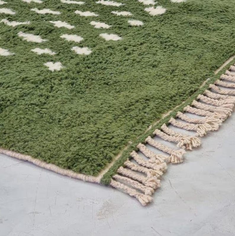 A plush handwoven Moroccan Berber rug in a rich moss green. The thick wool carpet features a solid dark green band near the top and a lighter mossy green base beautifully adorned with small, cream-colored geometric diamond motifs at the bottom. It is draped elegantly against a white painted brick wall next to a gold accent table with trailing ivy, perfectly capturing a warm, earthy, and organic modern interior design aesthetic.