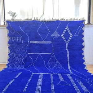 Striking handmade Moroccan rug in bold cobalt blue with white tribal Amazigh symbols. Crafted from natural wool, this unique Berber rug features traditional abstract lines and diamond motifs, perfect for adding a splash of color and culture to modern interiors. Ideal for boho, eclectic, or contemporary homes looking for a statement piece. Soft texture and timeless authenticity.