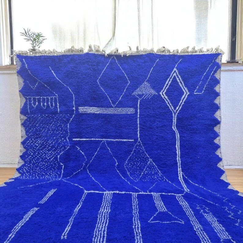 Striking handmade Moroccan rug in bold cobalt blue with white tribal Amazigh symbols. Crafted from natural wool, this unique Berber rug features traditional abstract lines and diamond motifs, perfect for adding a splash of color and culture to modern interiors. Ideal for boho, eclectic, or contemporary homes looking for a statement piece. Soft texture and timeless authenticity.