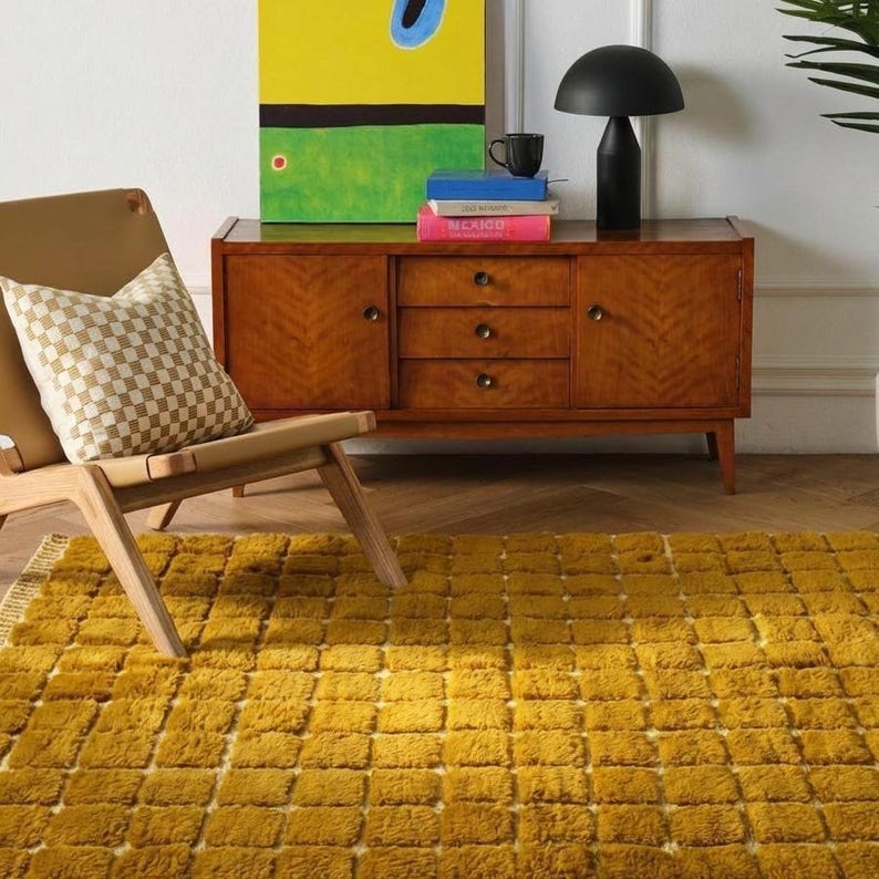 Large handmade Moroccan high-pile shag rug in saturated Thai curry mustard-gold, displayed vertically against a white brick wall. Surface features a tone-on-tone square grid pattern formed through pile variation — raised mustard squares separated by recessed ivory cream channel lines across the entire field. Dense, plush, curly wool pile texture throughout. Knotted braided ivory fringe along the top hem edge and long hand-tied ivory fringe along the bottom edge.